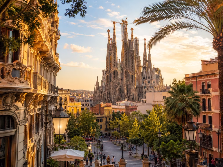 Golden-hour cityscape of Barcelona, Spain, with the Sagrada Família rising above palm-lined streets, historic buildings, greenery, and pedestrians in warm Mediterranean light.