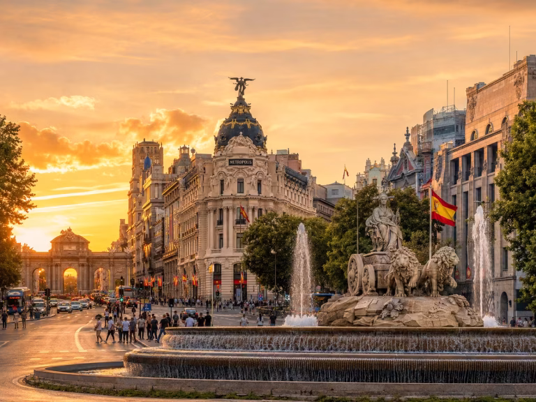 A golden-hour cityscape of Madrid featuring the Cibeles Fountain in the foreground, historic architecture along a busy boulevard, Spanish flags, pedestrians, and the Puerta de Alcalá in the distance.