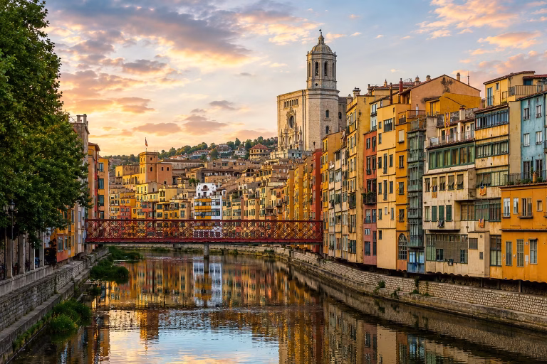 Scenic view of Girona, Spain, with colorful riverside buildings reflected in the Onyar River, a red iron bridge, Girona Cathedral rising above the old town, and a warm sunset sky.
