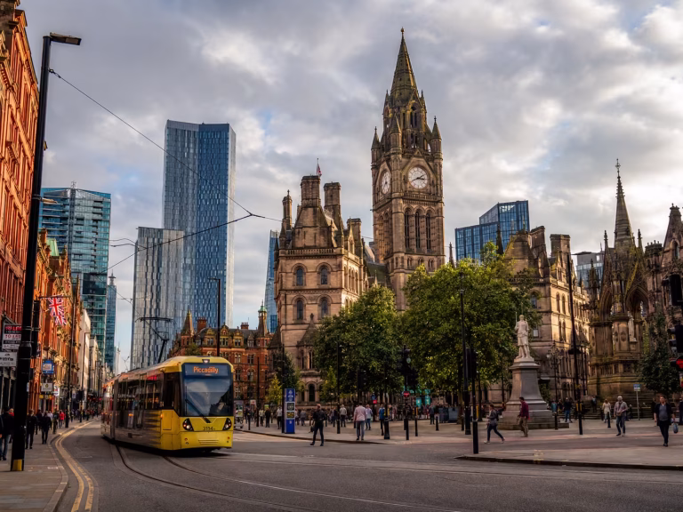 A Manchester city-centre street scene showing a yellow Metrolink tram, historic Gothic-style architecture, modern glass towers, pedestrians, trees, and a cloudy English sky.