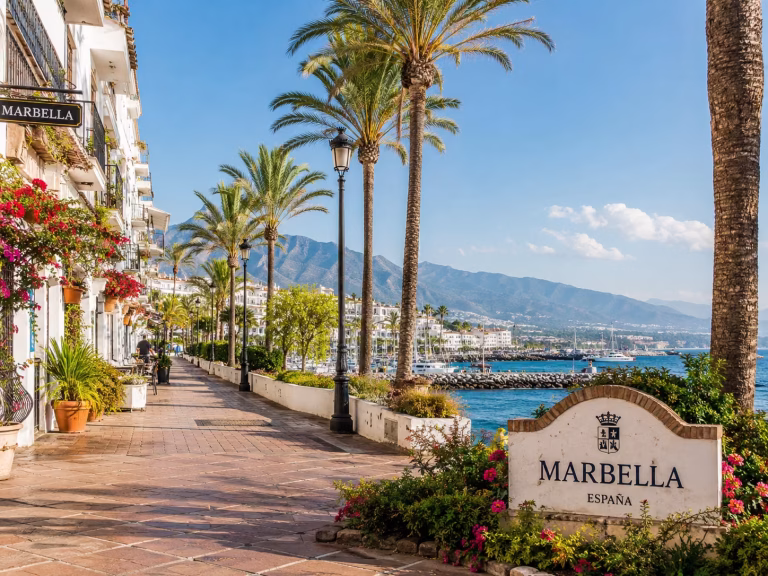 Sunlit coastal promenade in Marbella, Spain, with palm trees, whitewashed buildings, colorful flowers, a Marbella sign, blue Mediterranean water, marina views, and mountains in the distance.
