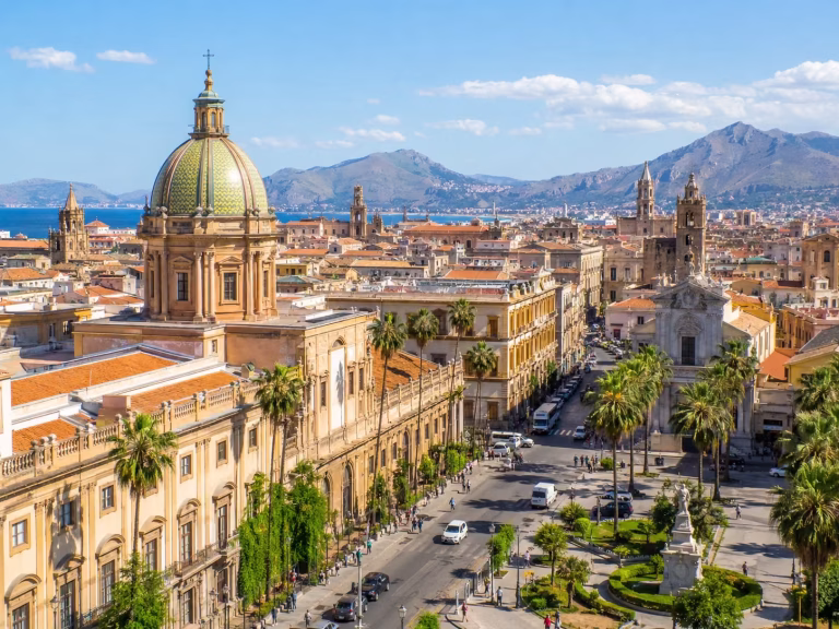 Sunny panoramic view of Palermo, Italy, with historic domes, church towers, palm-lined streets, terracotta rooftops, distant mountains, and the Mediterranean coastline in the background.